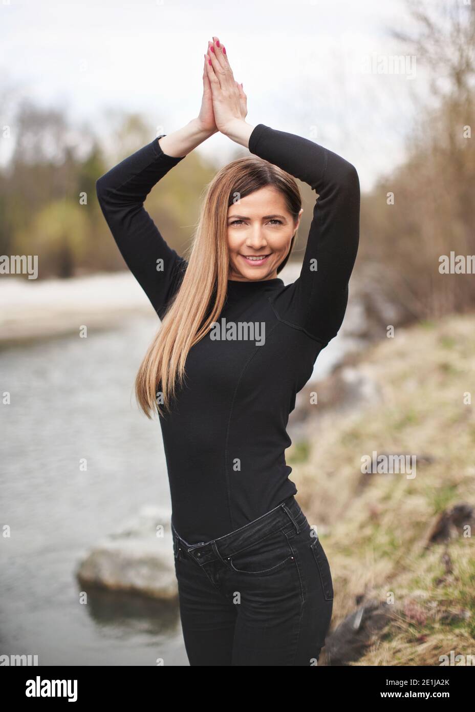 Giovane donna con capelli lunghi in cima nera che si esercita vicino al fiume, tenendo le mani sopra la testa Foto Stock