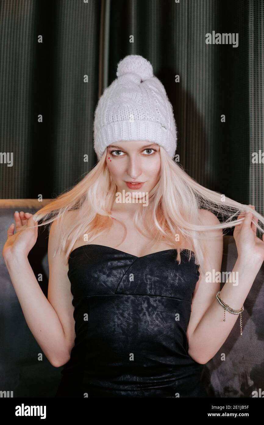 Bella ragazza bionda indossa un cappello a maglia calda con pompon e un elegante abito scuro senza strappo; lei sta tirando i capelli ai lati con entrambe le mani Foto Stock