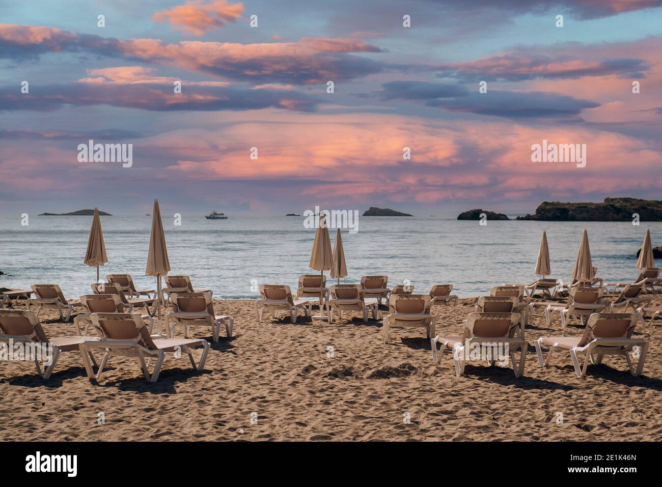 Ombrelloni ripiegati e sedie a sdraio vuote sulla spiaggia di sabbia di Ibiza durante multicolore pittoresco tramonto sera luce romantica cielo vista, Isole Baleari Foto Stock