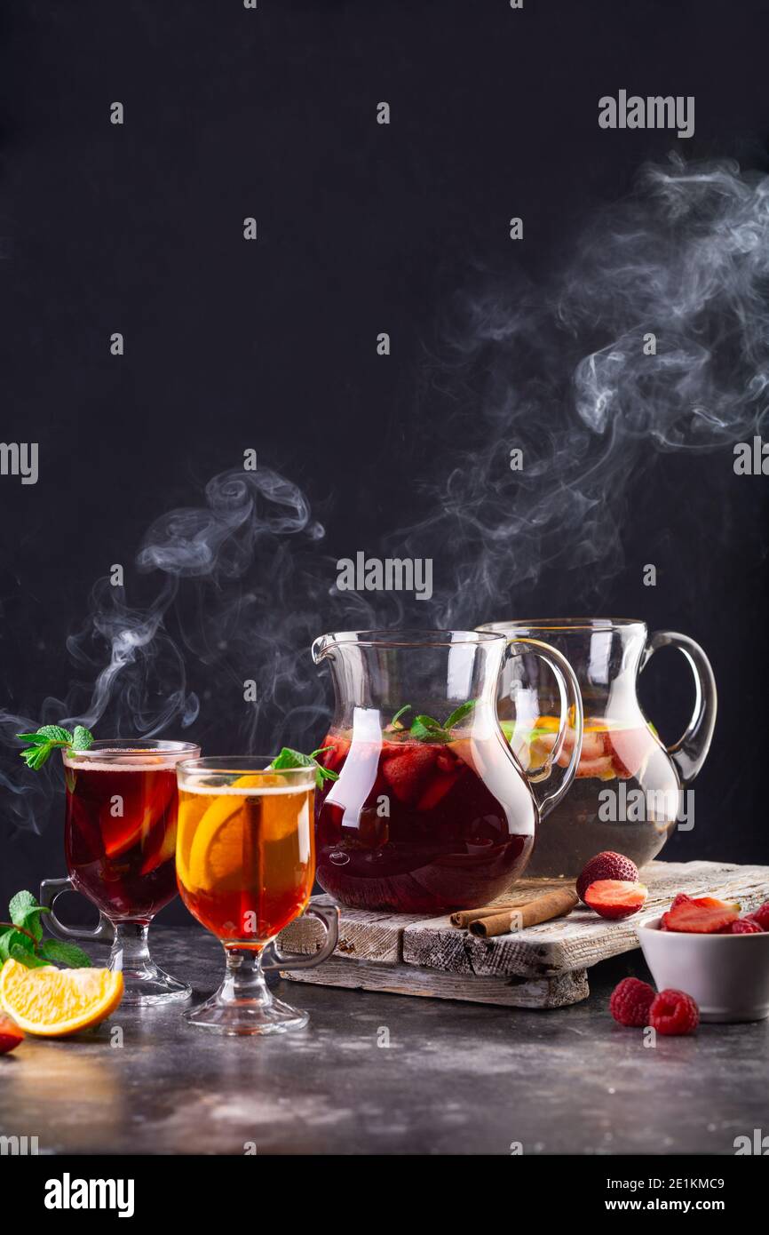 Sul tavolo, due brocche con limonata, VIN brulé e bicchieri pieni di bevande. Il vapore si sviluppa dalle bevande. Fragole, apeltsin, lamponi e. Foto Stock