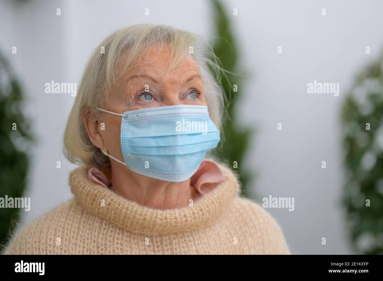 Donna anziana dagli occhi blu che indossa una maschera facciale come protezione durante La pandemia di Covid-19 in piedi all'aperto guardando in su con un attento espressione in a. Foto Stock