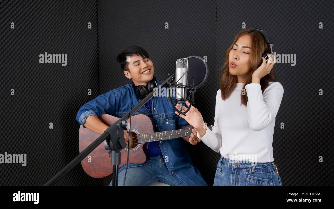 Giovane donna asiatica che canta di fronte alla parete insonorizzata nera. Il suo ragazzo suonò la chitarra insieme. Musicisti che producono musica in re professionale Foto Stock