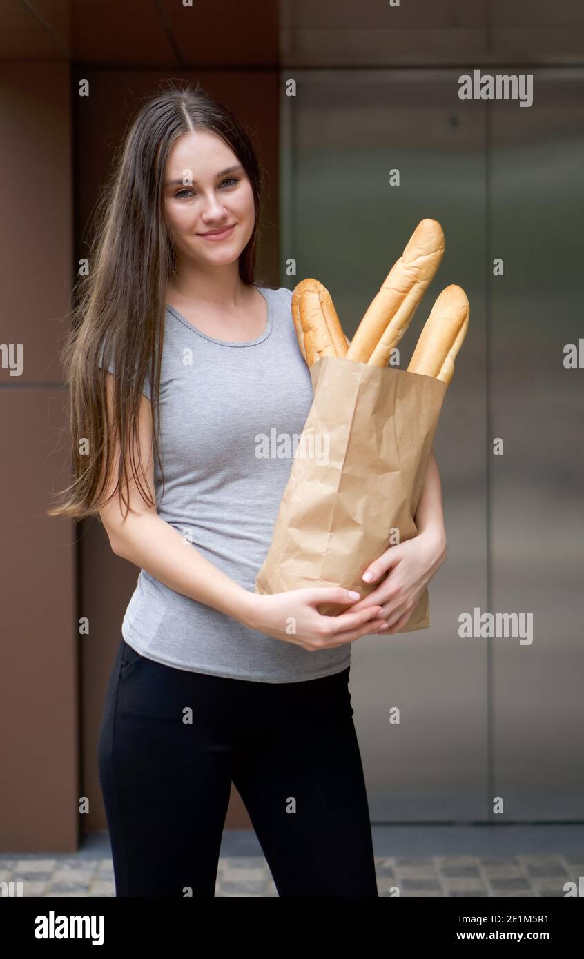 Giovane donna caucasica in abbigliamento casual che porta una borsa di carta con baguette, stand sorridente di fronte all'ascensore condominio. Cibo e pacchi deli Foto Stock