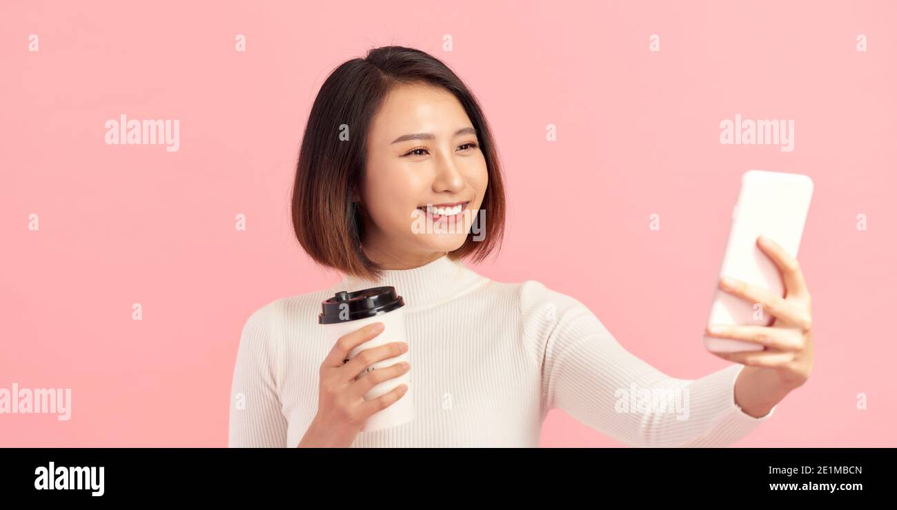 Ritratto di una donna sorridente attraente che prende un selfie mentre tenendo la tazza di caffè take away su sfondo rosa Foto Stock