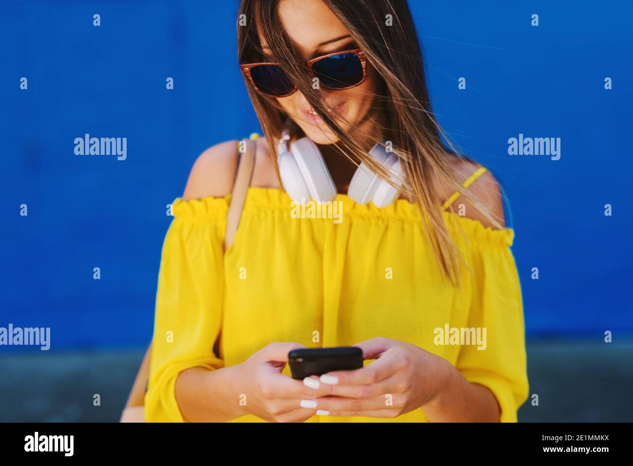 Ragazza carina in camicia gialla che si trova di fronte alla parete blu e tenendo cellulare con sorriso sul viso. Foto Stock