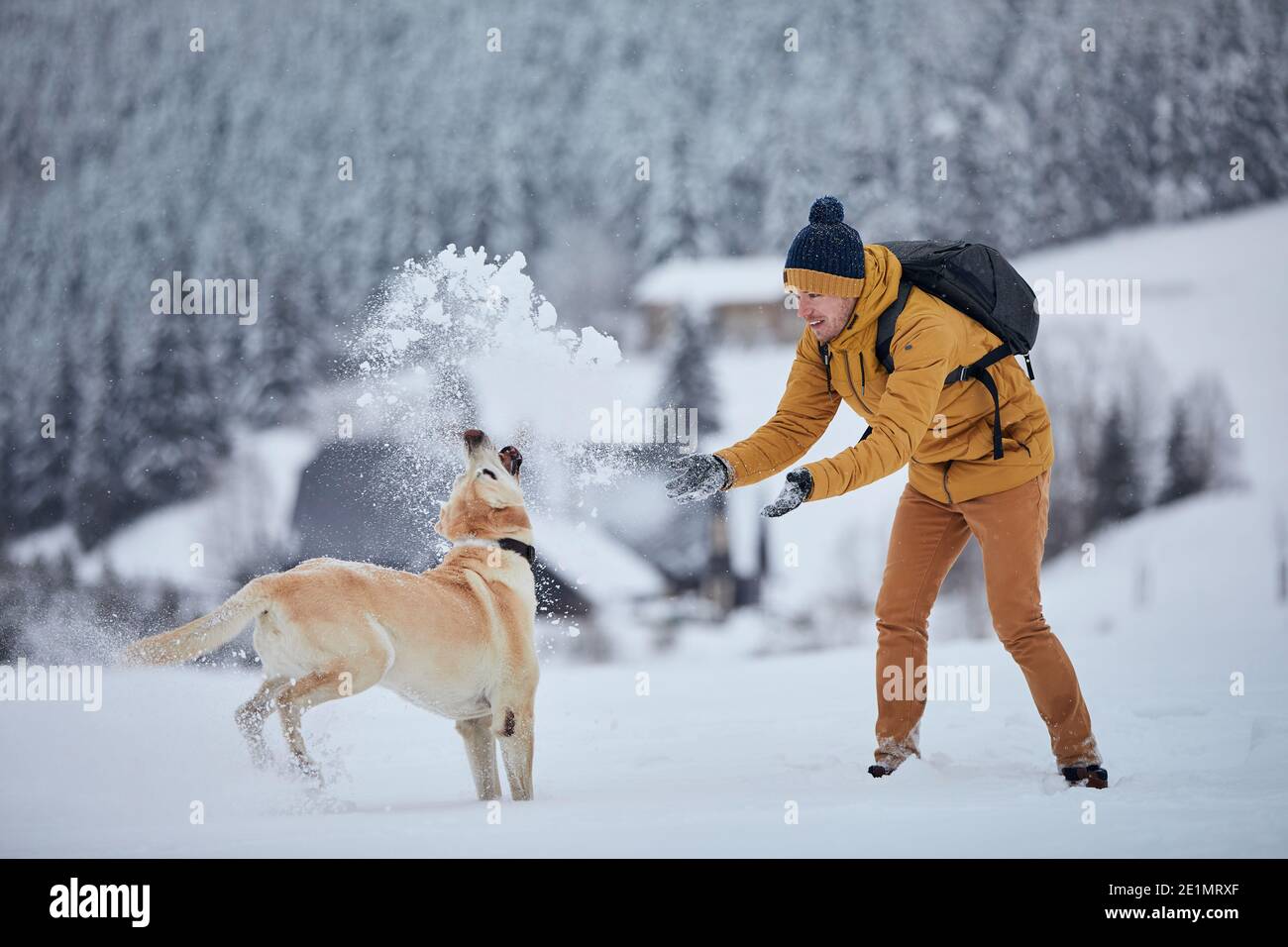Giovane uomo con cane in inverno. Proprietario di animali domestici con il suo labrador Retriever giocare nella neve contro il vecchio villaggio in bella natura. Montagne Jizera, Repubblica Ceca Foto Stock
