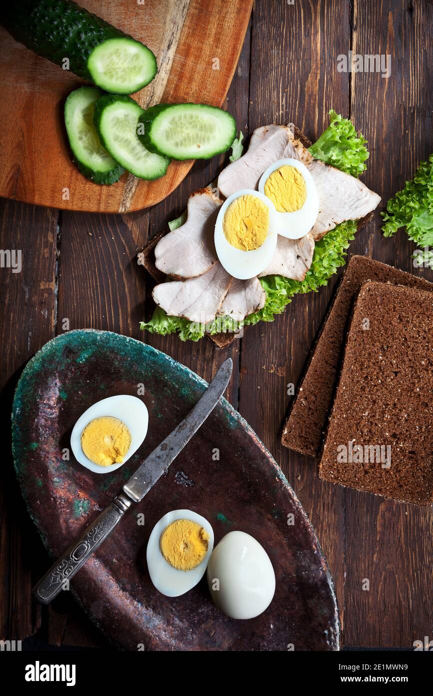 preparare sandwich di carne con foglie di lattuga verde, uova tritate, cetrioli e pane di segala o grano, su sfondo di legno rustico scuro, nessuno Foto Stock