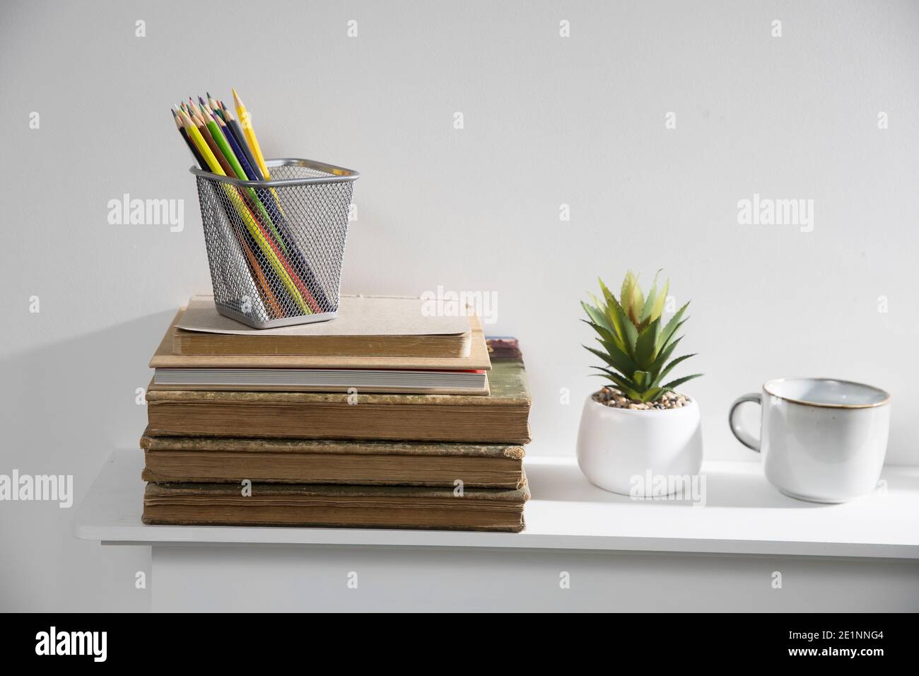 Portamatite con pastelli su pila di vecchi libri d'epoca sdraiati su mantelpiece. Aloe artificiale in vaso di ceramica e tazza di porcellana. Spazio di copia Foto Stock