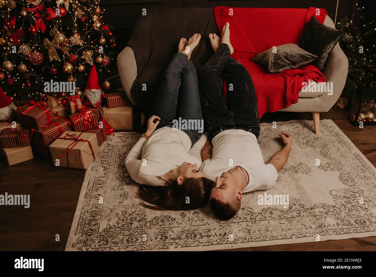 Giovane coppia uomo e donna si trovano sul pavimento vicino divano con gambe piedi su baciare abbracciando nella camera da letto vicino albero di natale. Casa decorata per Capodanno. Foto Stock