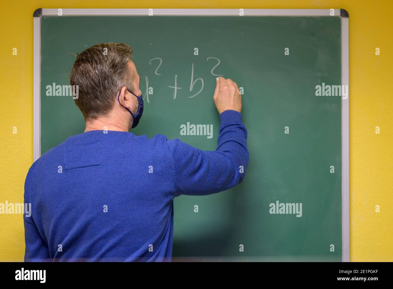 Insegnante che indossa una maschera facciale durante la pandemia del coronavirus in piedi con la sua schiena allo spettatore che scrive un'equazione matematica su una lavagna Foto Stock