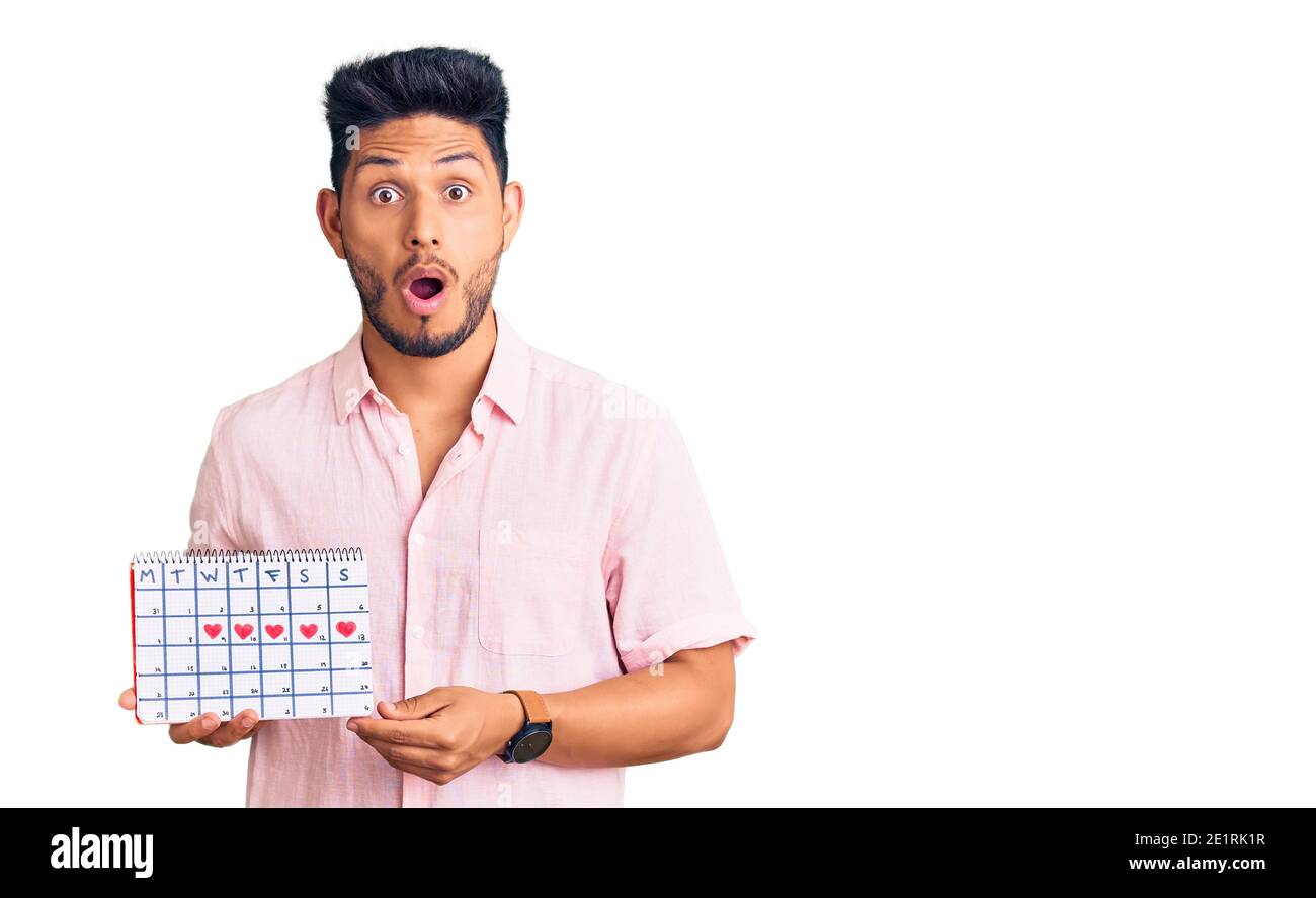 Bell'uomo latino-americano giovane tenendo cuore calendario spaventato e stupito con bocca aperta per sorpresa, viso incredulo Foto Stock