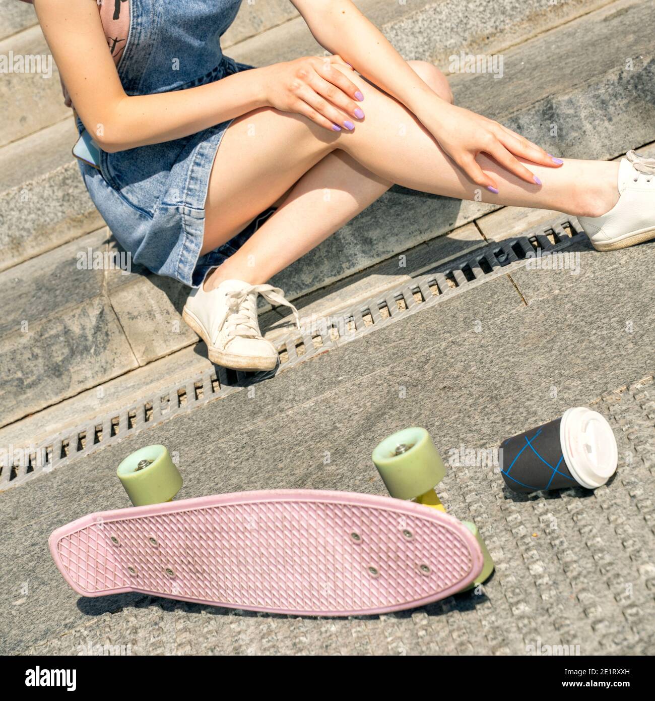 una ragazza in denim si siede sui gradini di pietra con uno skateboard e una tazza di caffè. danni al ginocchio. sport pericolosi in città Foto Stock