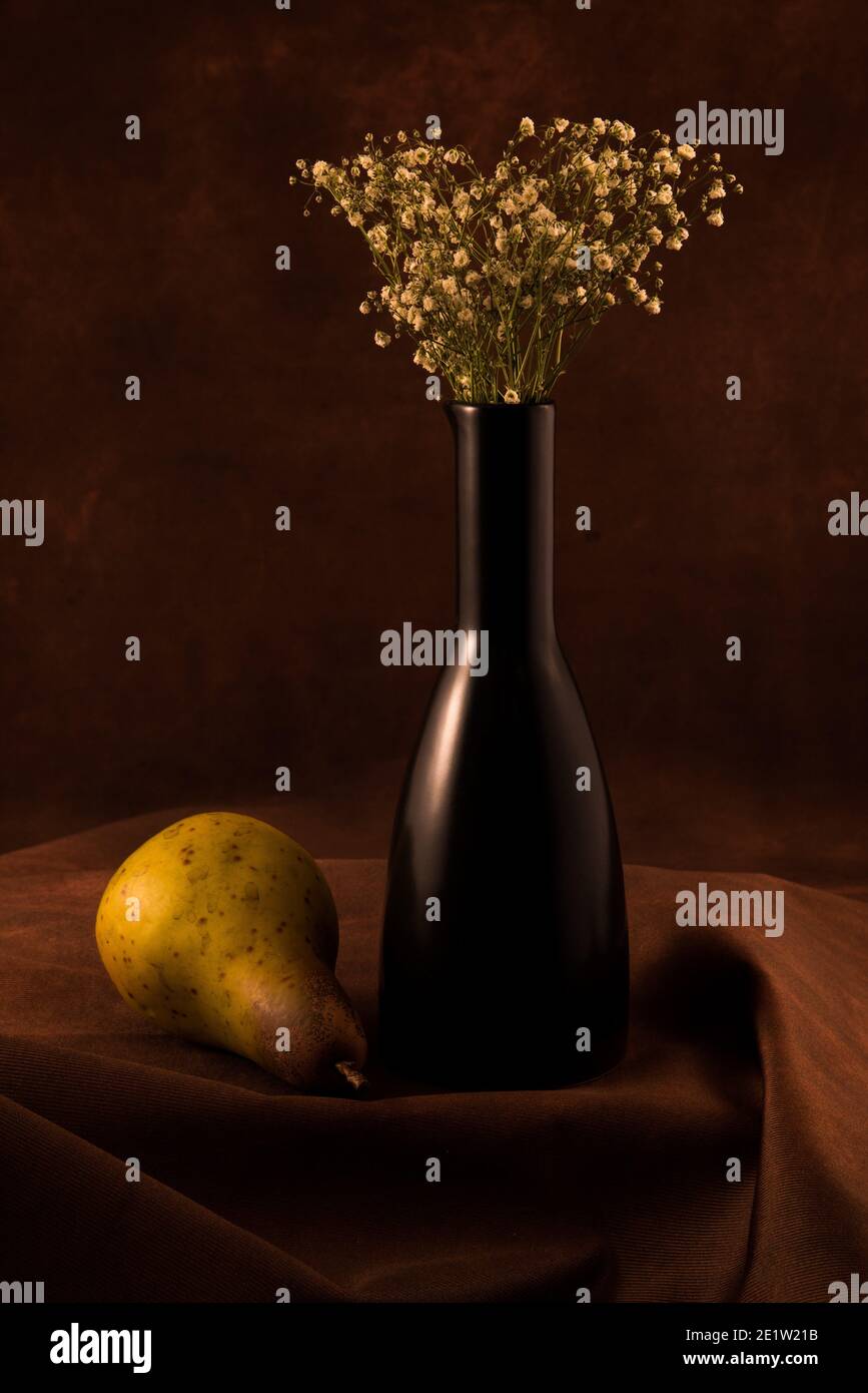 Vaso nero singolo con fiori di alito 'baby's', a contrasto con le pere. Una vita still vintage con illuminazione soffusa. Foto Stock