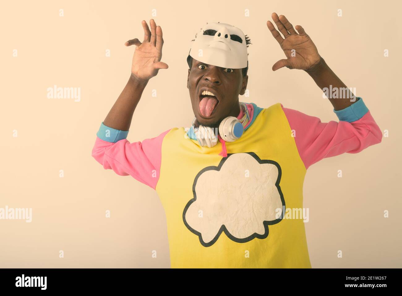Studio di giovane uomo africano nero che stacca la lingua fuori indossando una maschera bianca sulla testa e sulle cuffie intorno al collo con entrambe le braccia sollevate Foto Stock