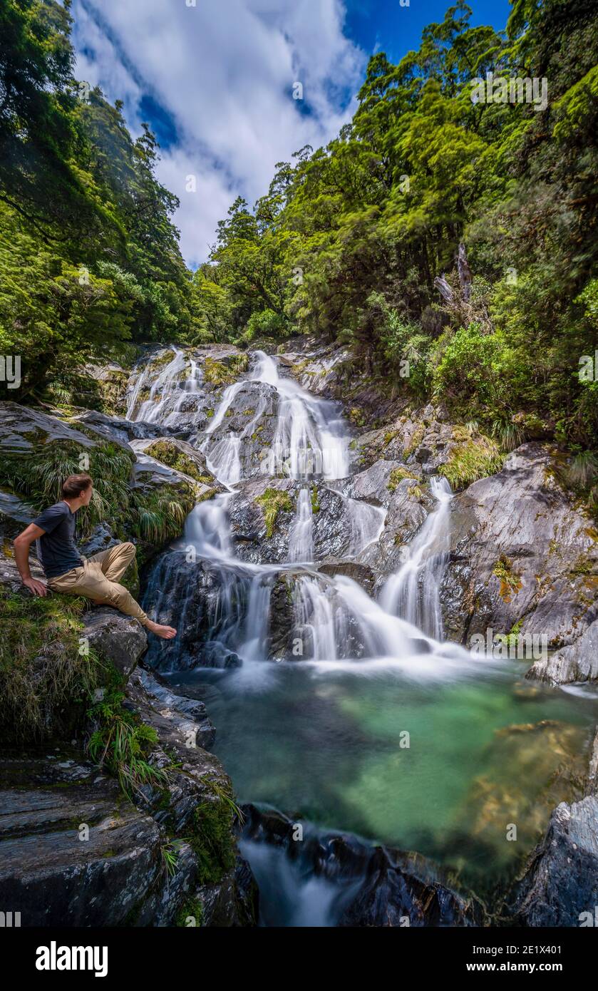 Giovane uomo seduto a una cascata, Fantail Falls, Makarora River, Wanaka, West Coast, South Island, Nuova Zelanda Foto Stock
