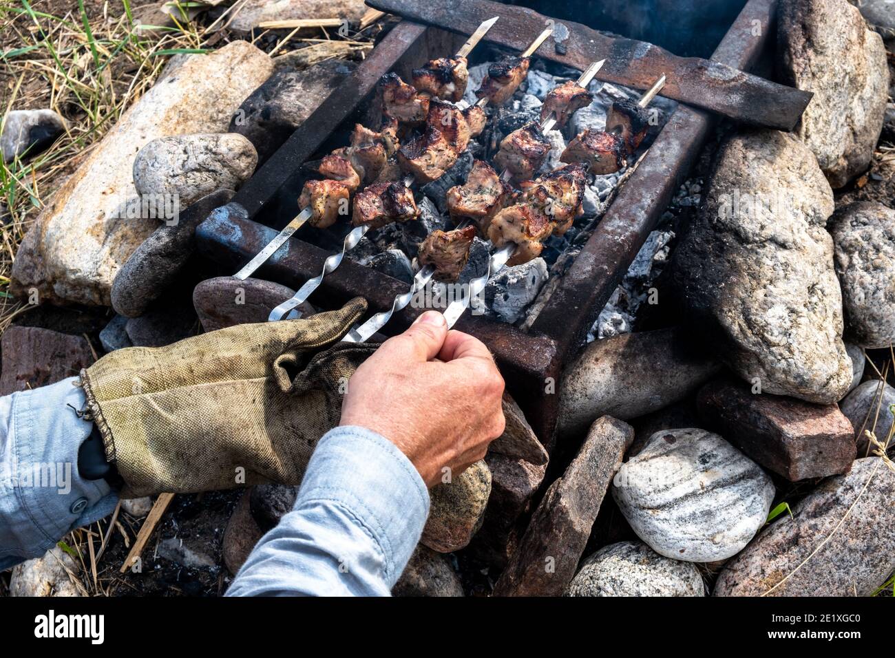 Deliziosa e appetitosa carne alla griglia, cotta al fuoco nel cortile di una casa di campagna. Le mani maschili prendono gli spiedini e li girano per cucinare meglio. Foto Stock