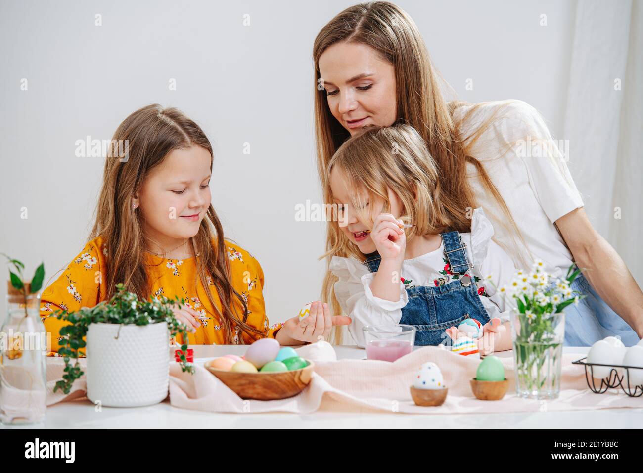 Madre amorevole che osserva le sue due figlie mentre dipingono le uova di pasqua dietro la tabella Foto Stock