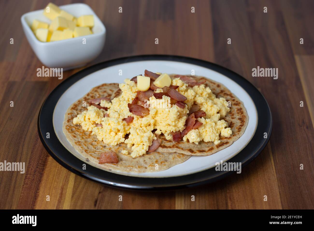 Una vista angolata delle torte di oat del Derbyshire servite con uova strapazzate e pancetta con un piatto di burro leggermente fuori fuoco sullo sfondo Foto Stock