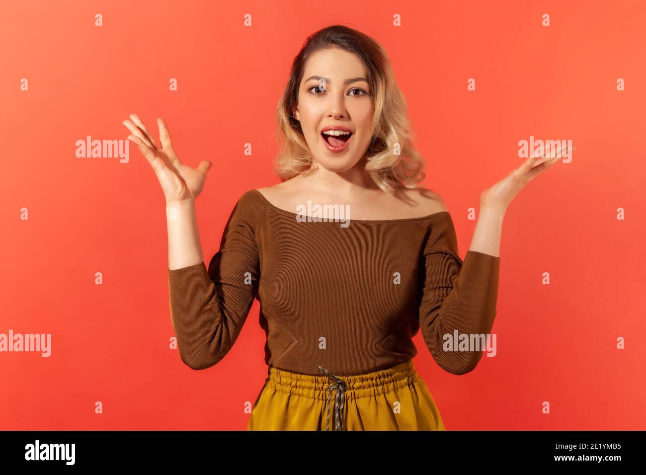 Ritratto di bella donna stupita bella con i capelli biondi in blusa marrone in piedi sorpresi con bocca aperta, le mani sollevate e guardando la macchina fotografica. Foto Stock