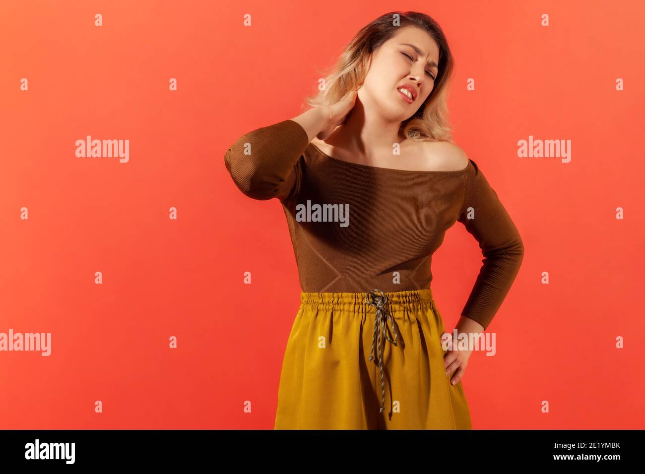 Dolore al collo o dolore. Ritratto di bella blonde giovane donna adulta in blusa marrone in piedi e tenendo il suo collo doloroso. Studio interno con isol di ripresa Foto Stock