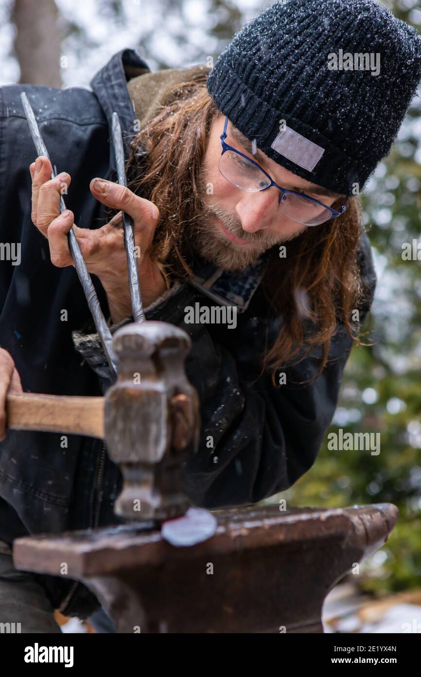 Primo piano di un giovane fabbro sotto la neve, concentrato nel martellare e appiattire una rossa, sfrigolante asta di ferro calda fuori dalla fucina. Foto Stock