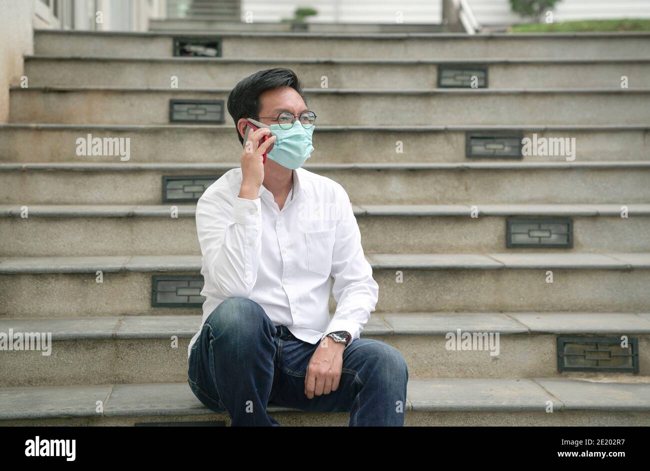 Uomo d'affari asiatico che indossa la maschera di faccia seduto su scala, utilizzando e parlando sul suo telefono. Foto Stock