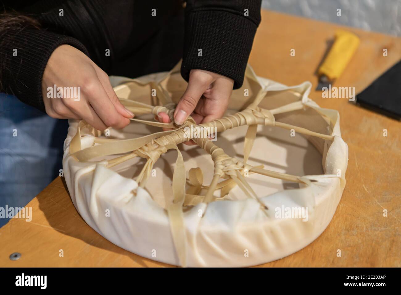 Istantanee da un laboratorio di drum nativo. Primo piano delle mani di una giovane donna che lavora un pezzo di pelle intorno a un supporto in legno per creare un tamburello. Foto Stock