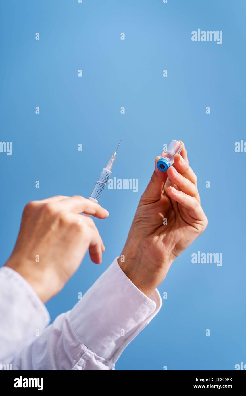 Primo piano sulla mano di sconosciuta donna caucasica medico tenuta siringa con ago e ampolla di vaccino di fronte parete blu - covid-19 coronavir Foto Stock