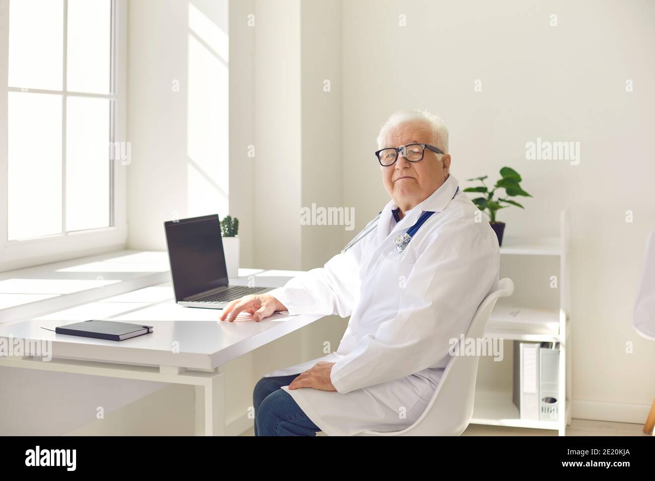Medico professionista senior positivo in uniforme seduto con il computer portatile in ufficio medico e guardando la fotocamera Foto Stock