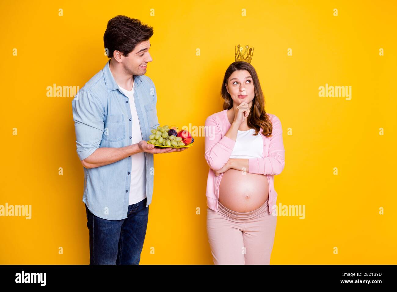 Foto di giovane coppia in piedi donna incinta pensare indossare corona ragazzo con frutta isolato su sfondo giallo Foto Stock