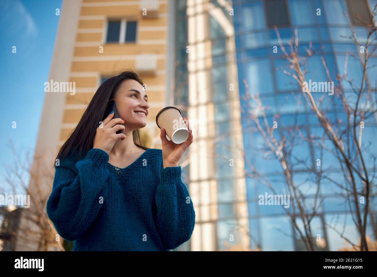 Bella donna allegra guardando da parte con un sorriso lucido tenendo tazza di bevanda calda in mano e ascoltare qualcuno dal suo smartphone Foto Stock