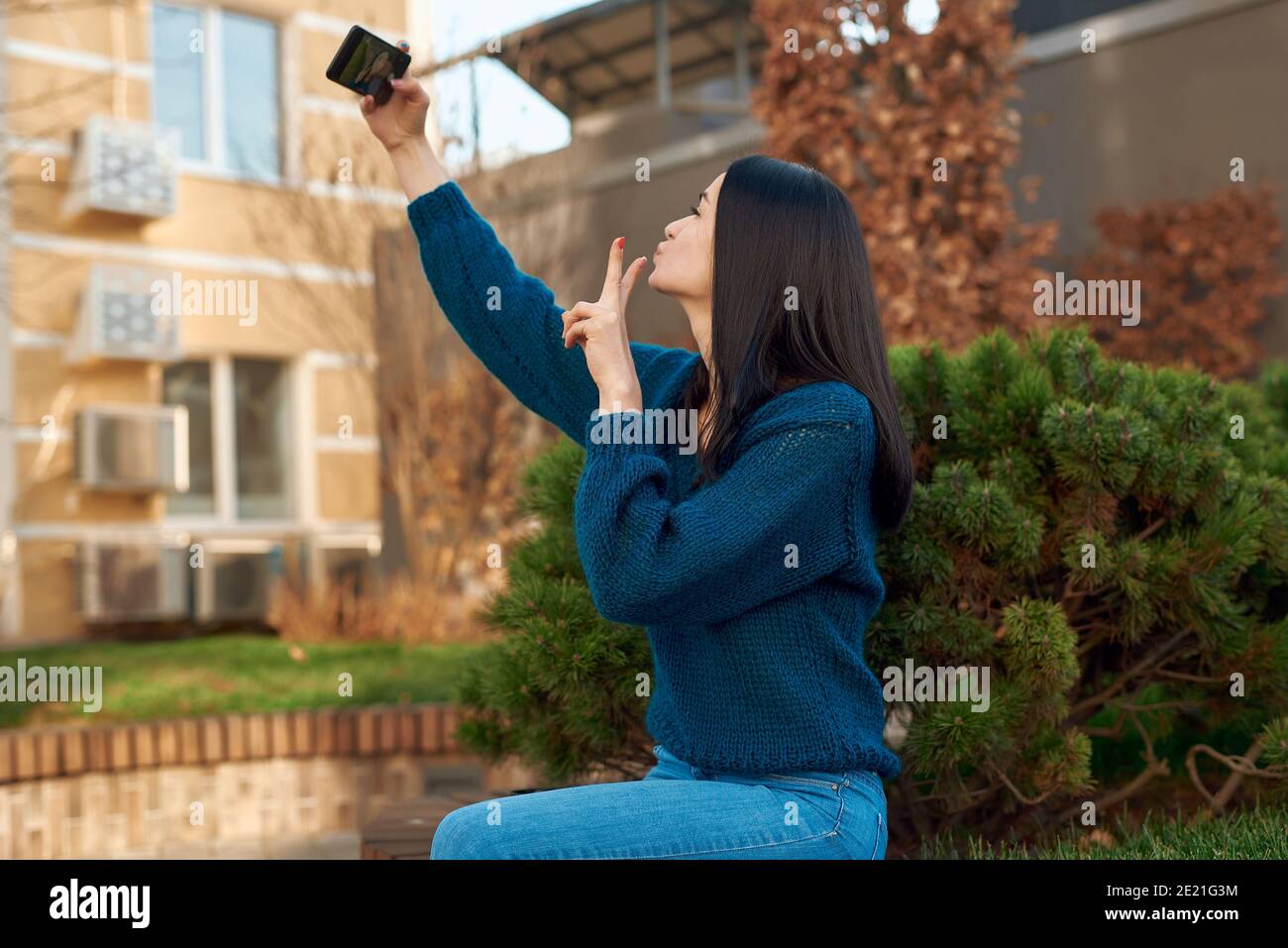 Vista laterale di una bella studentessa che si prende foto all'aperto con una piccola conifera soffice sullo sfondo Foto Stock