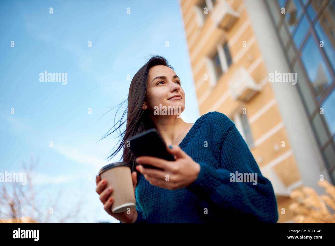 Giovane bella donna passeggiando lungo alti edifici moderni e tenuta telefono cellulare e tazza di caffè in mani Foto Stock