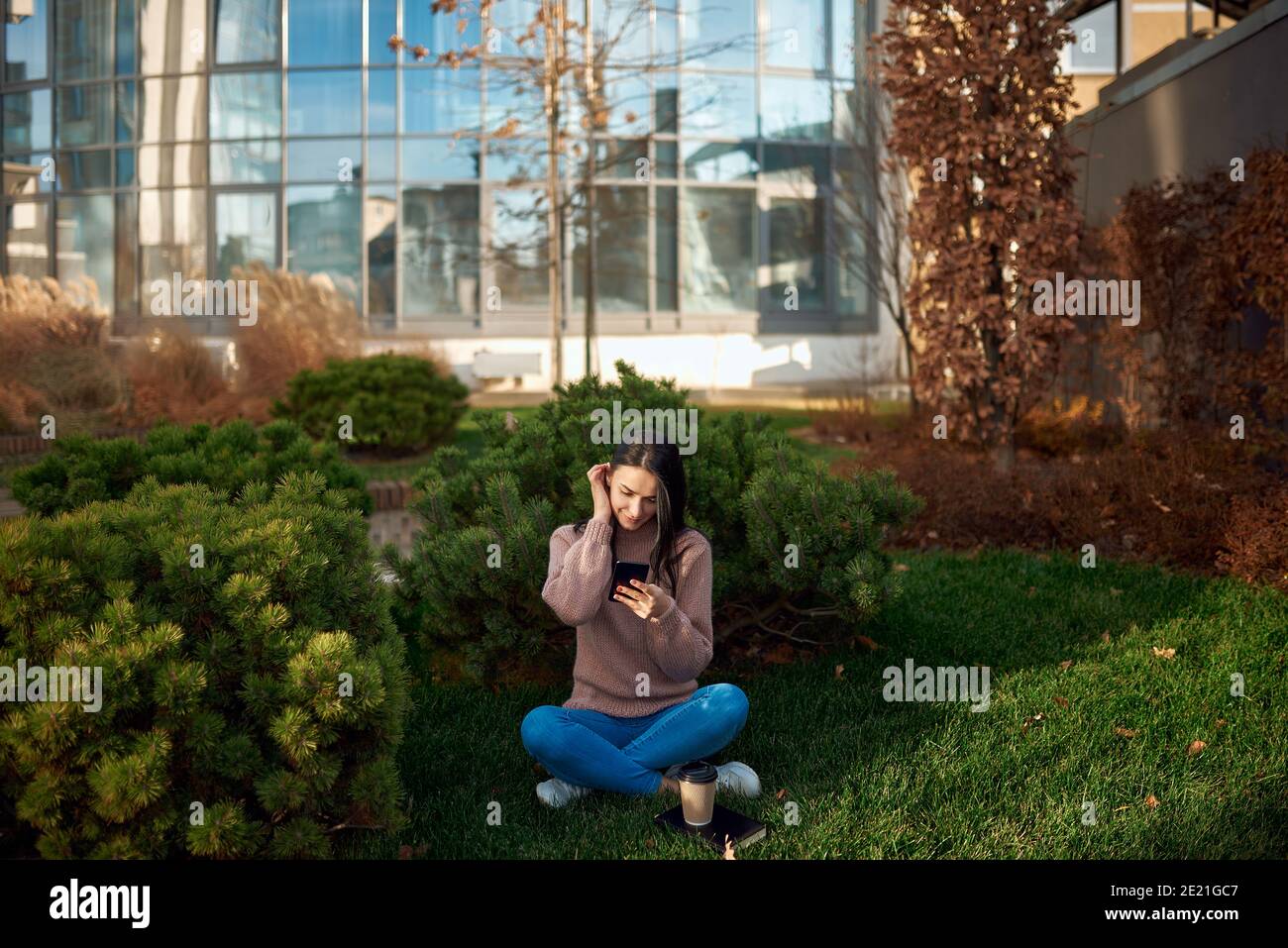 Giovane donna pensiva che guarda sullo schermo del suo telefono cellulare e seduto su erba con alto edificio sullo sfondo Foto Stock