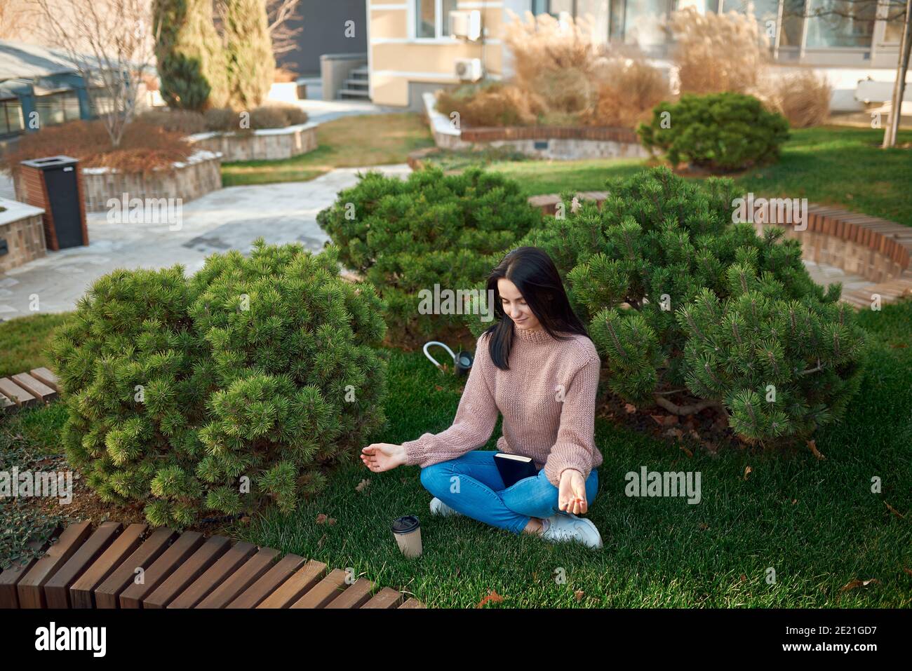 Vista ad alto angolo di una piacevole giovane femmina in cui rilassarsi una posa meditativa su un'erba in cortile accogliente dentro di fronte a edifici moderni Foto Stock