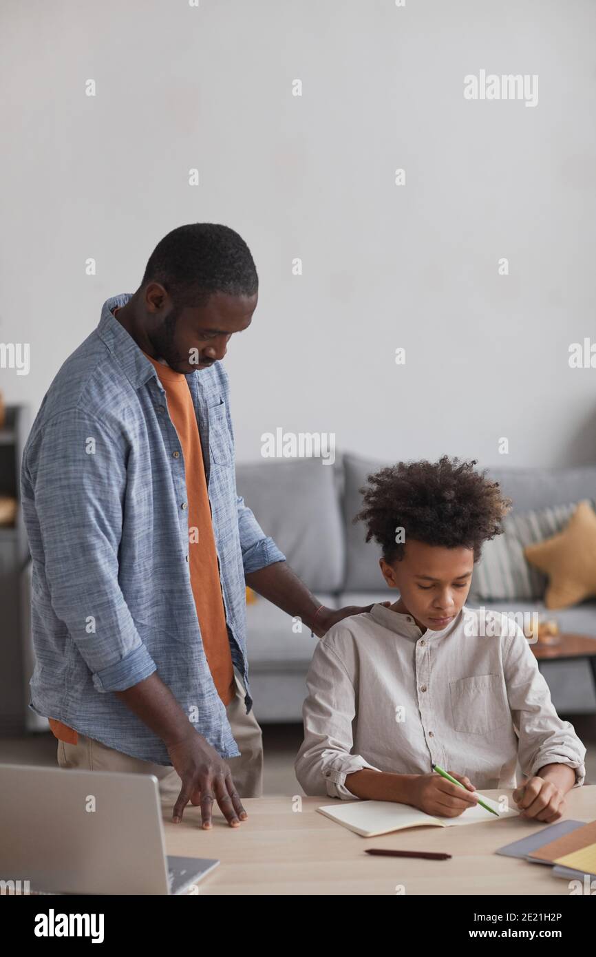 Ritratto verticale del padre afroamericano che aiuta il figlio che fa i compiti alla scrivania nell'interno domestico minimo, spazio di copia sopra Foto Stock