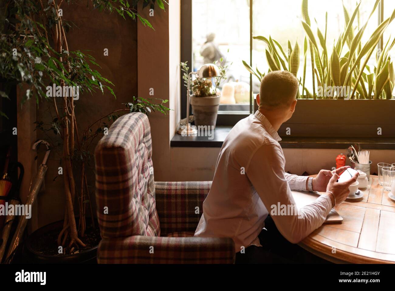 Giovane uomo d'affari che guarda fuori la finestra nel bar e tiene il suo telefono cellulare. Concetto di stile di vita Foto Stock