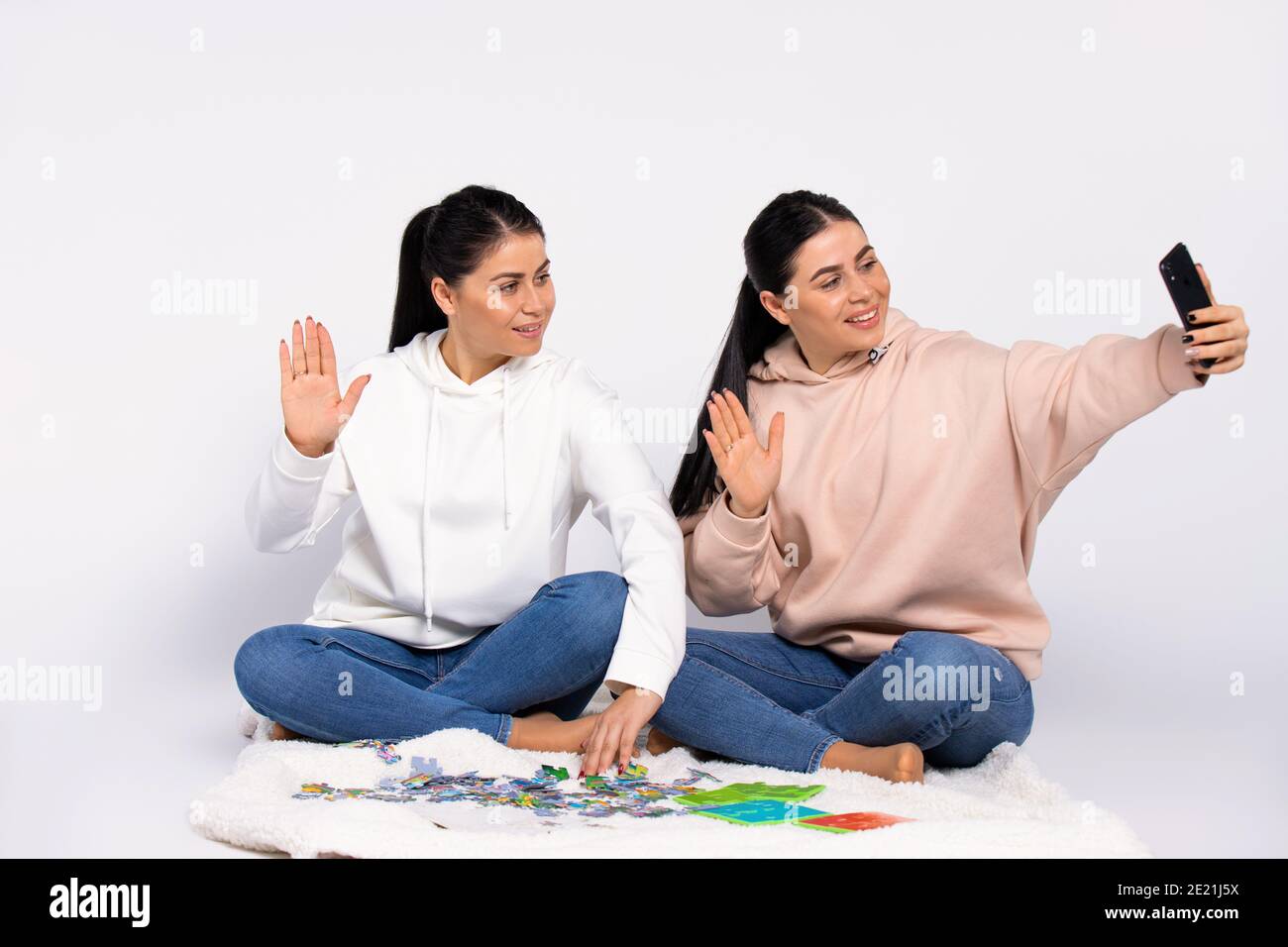I gemelli sono seduti sul pavimento, mettendo insieme un puzzle e comunicando tramite videochiamata su uno smartphone. Sfondo bianco. Foto Stock