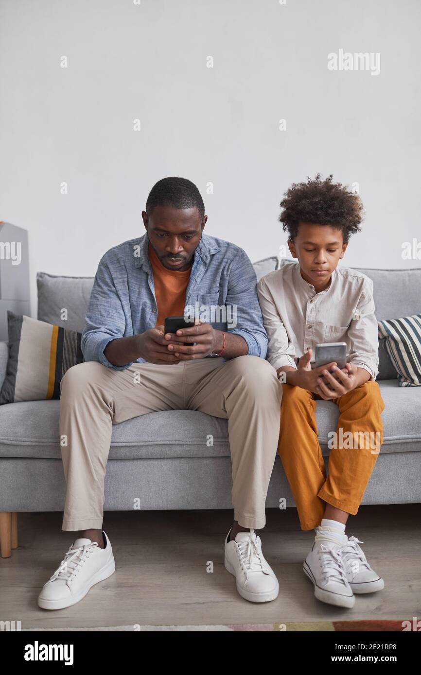 Immagine verticale di un ragazzo teenager afro-americano di stile che passa del tempo con il padre a casa seduto sul divano e giocando videogiochi sugli smartphone Foto Stock