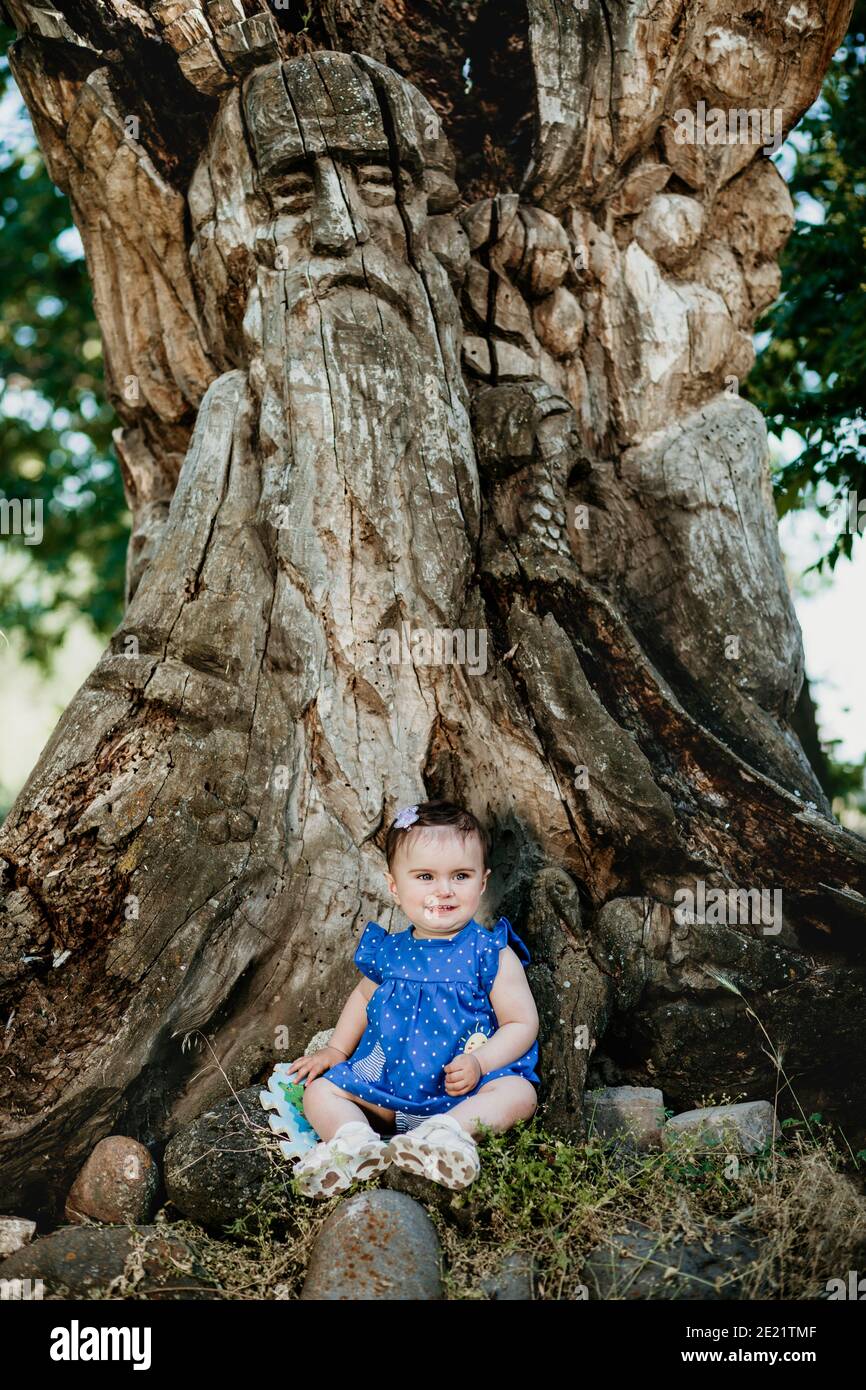 Bambino alla moda con abito blu seduto al vecchio albero gigante e sorridendo Foto Stock
