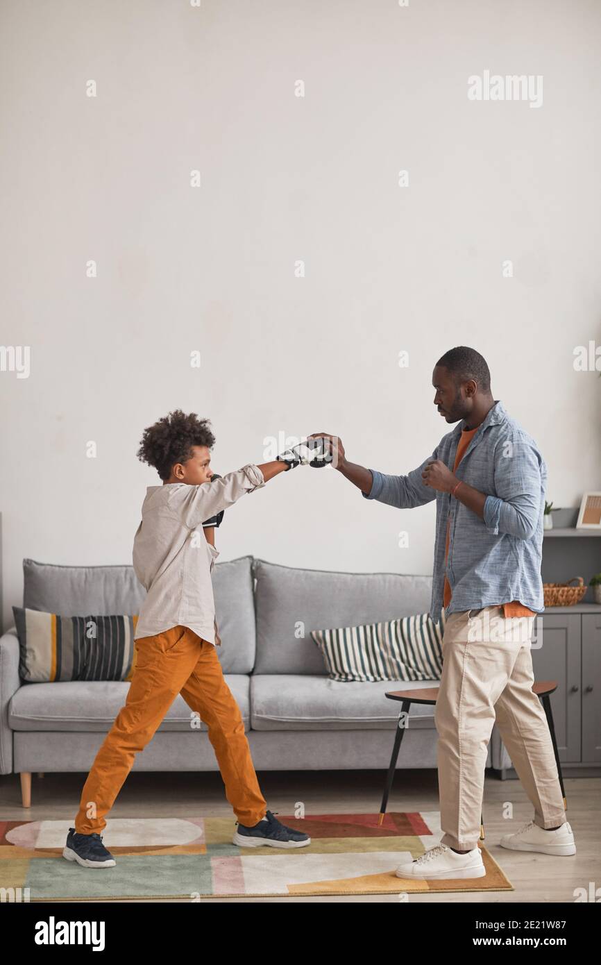Moderno padre afroamericano che pratica pugilato pugni con il suo figlio teen in soggiorno a casa, verticale vista laterale lungo colpo Foto Stock