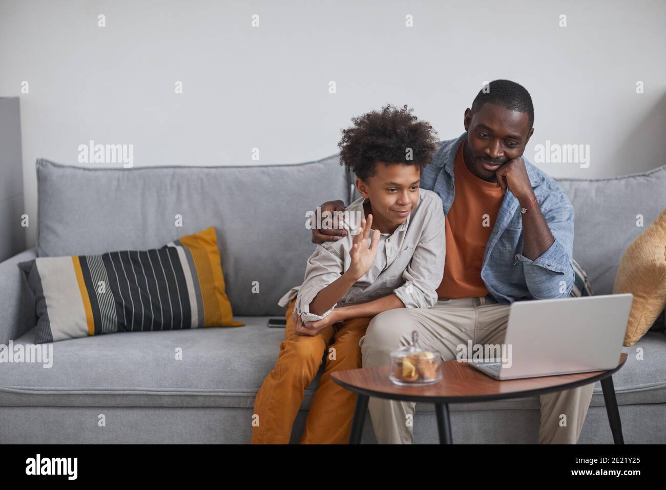 Padre e figlio moderni seduti sul divano di fronte schermo del portatile che avvia la videochiamata con i parenti che li salutano Foto Stock