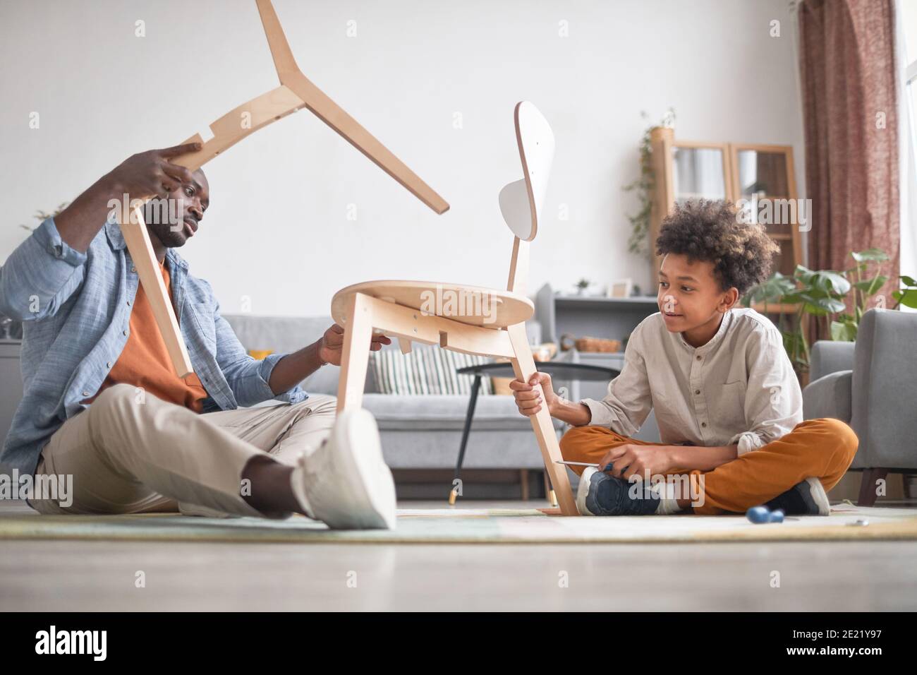 L'uomo di famiglia afroamericano che passa il tempo con il suo figlio teen in soggiorno a casa fissaggio sedia rotta Foto Stock