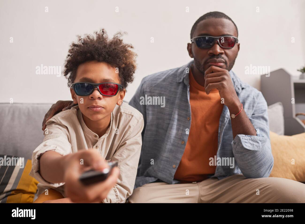 Ritratto medio di un elegante ragazzo afro-americano seduto sul divano Con suo padre che accende la pellicola 3D usando il clicker Foto Stock