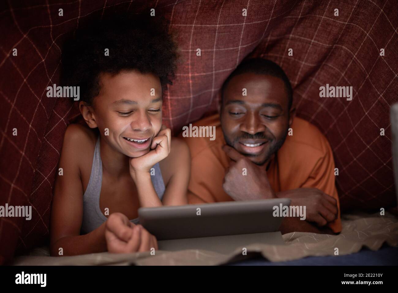 Gioioso ragazzo afroamericano guardando film emozionante su tablet digitale con suo padre sotto coperta controllata nelle tenebre Foto Stock