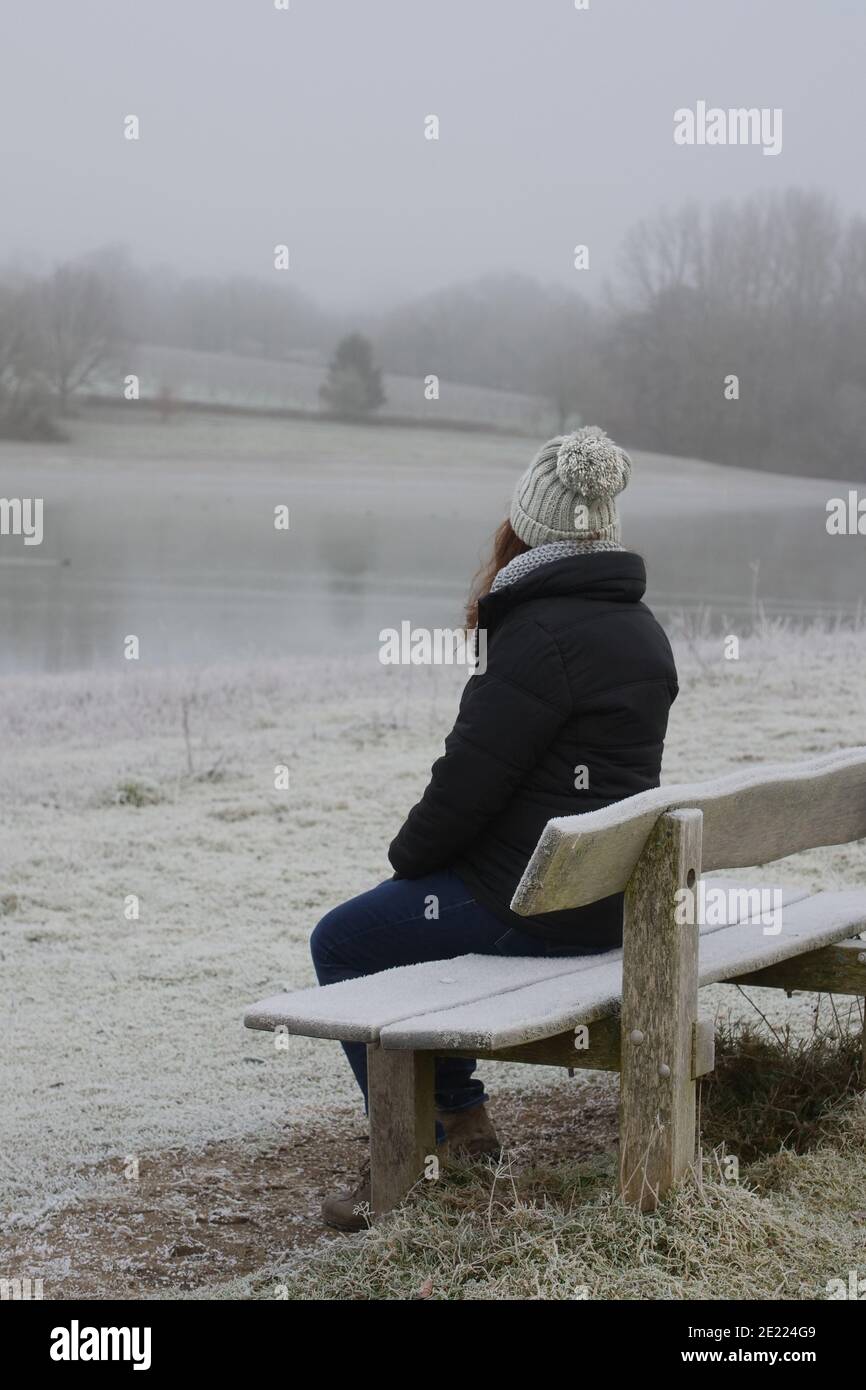 Una donna seduta indietro alla macchina fotografica su una panca di legno coperta dal gelo che si affaccia su un lago in inverno. Spazio di copia. Foto Stock