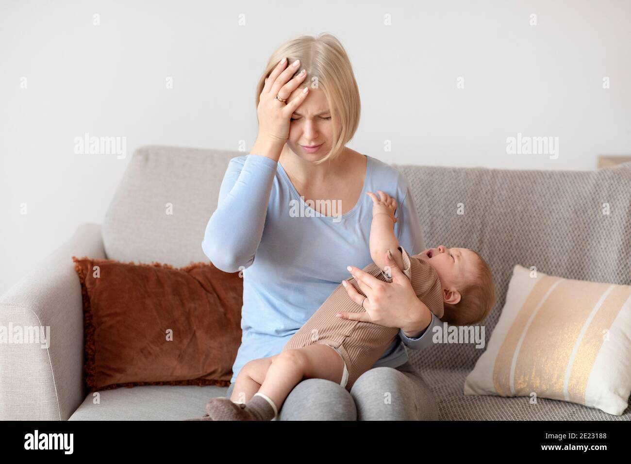 Donna bionda esausta che ha emicrania, tenendo piccolo bambino, interno domestico Foto Stock