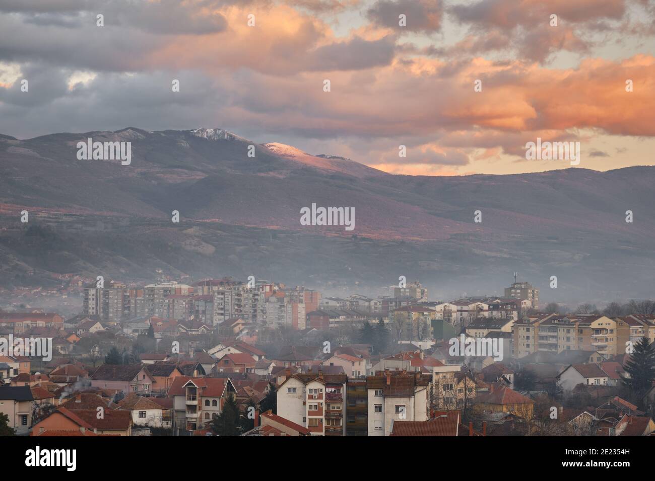 Misty, fumoso paesaggio urbano di Pirot durante il tramonto con impressionante, picco roccioso schiarita dalla luce dorata sullo sfondo e un cielo nuvoloso Foto Stock