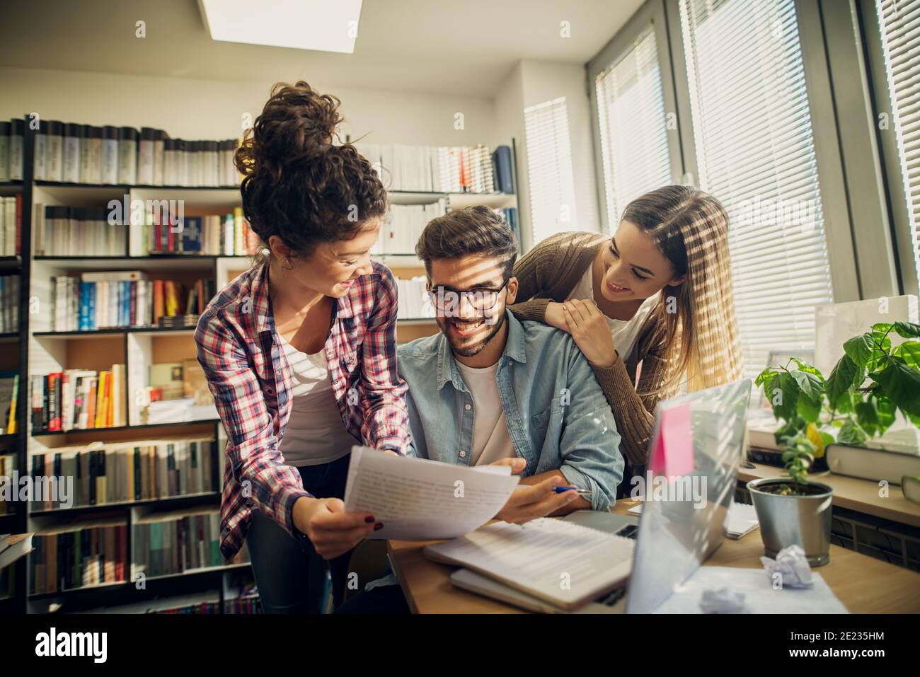 Due ragazze allietate allegre che aiutano il loro amico a passare exam.sitting in una biblioteca luminosa. Foto Stock