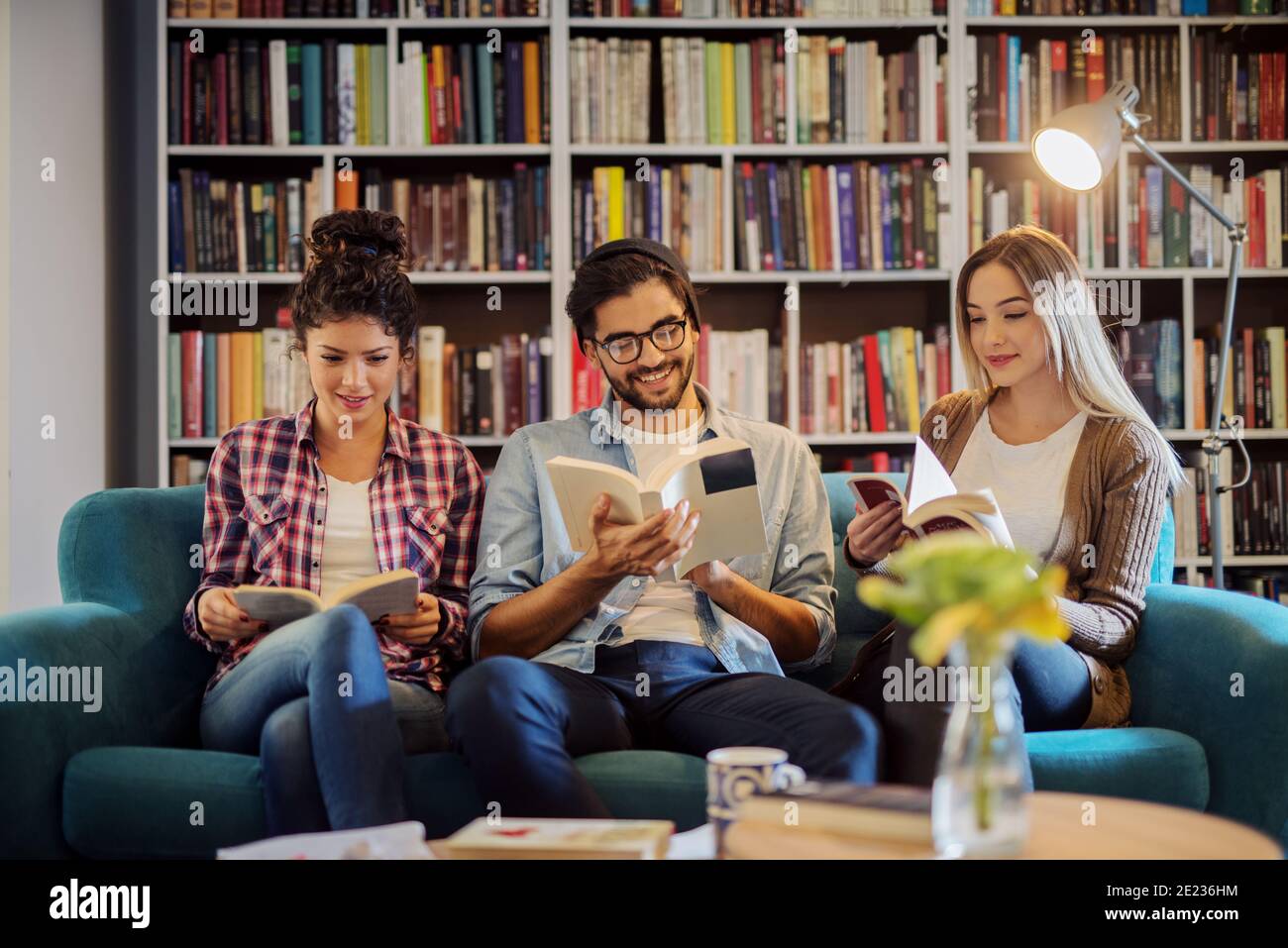 Tre giovani studenti moderni seduti in una moderna biblioteca e leggendo libri. Preparazione degli esami insieme. Foto Stock